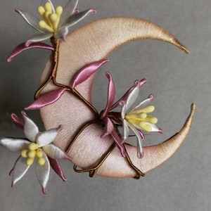 May include: A crescent moon-shaped decorative item with embroidered details. The moon is a soft pink color with gold-colored edges. Delicate white and pink flowers with yellow centers and brown stems are attached to the moon.