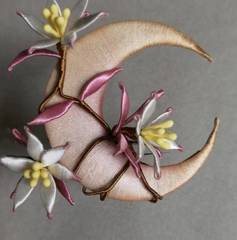 May include: A crescent moon-shaped decorative item with embroidered details. The moon is a soft pink color with gold-colored edges. Delicate white and pink flowers with yellow centers and brown stems are attached to the moon.