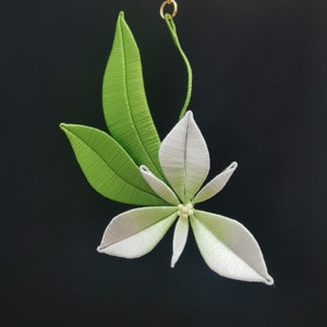 May include: A white silk flower with green leaves, hanging from a gold chain. The flower has five petals and a centre with small white beads.