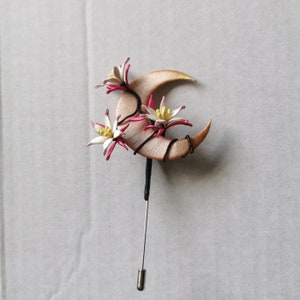 May include: A handcrafted brooch featuring a crescent moon design with three stylized flowers. The moon is a light tan color, and the flowers have white petals with pink and yellow accents. The brooch has a long pin with a safety clasp.