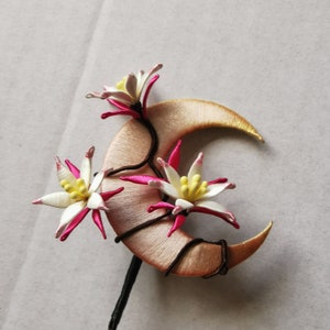 May include: A handcrafted hair accessory featuring a crescent moon shape in a soft peach color, adorned with white and pink floral accents. The flowers have yellow centers and are attached with dark brown wire. The stem is black.