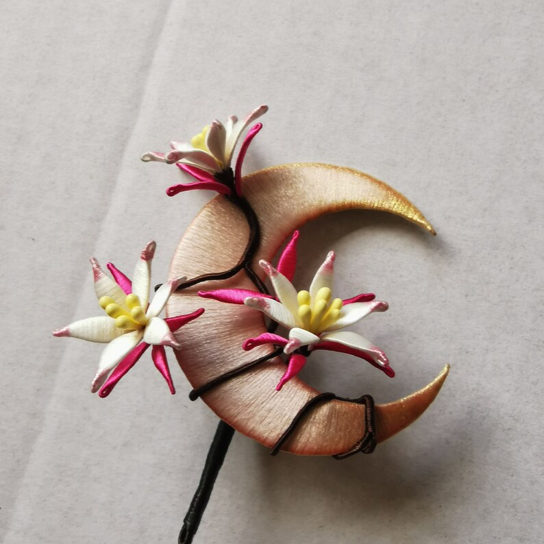 May include: A handcrafted hair accessory featuring a crescent moon shape in a soft peach color, adorned with white and pink floral accents. The flowers have yellow centers and are attached with dark brown wire. The stem is black.