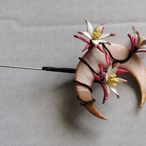 May include: A handcrafted lapel pin featuring a crescent-shaped design with floral accents. The pin has a silver-colored needle and a black base. The flowers are white with pink and yellow details.