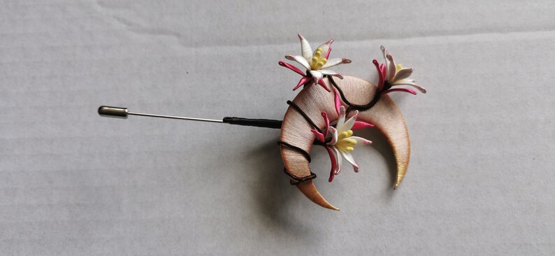 May include: A handcrafted lapel pin featuring a crescent-shaped design with floral accents. The pin has a silver-colored needle and a black base. The flowers are white with pink and yellow details.