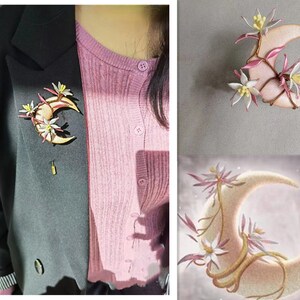 May include: A crescent moon brooch with floral accents. The brooch features a light beige moon shape with delicate pink and white flowers and vines. The brooch is pinned to a black blazer.