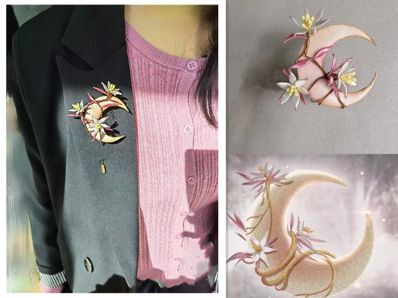 May include: A crescent moon brooch with floral accents. The brooch features a light beige moon shape with delicate pink and white flowers and vines. The brooch is pinned to a black blazer.