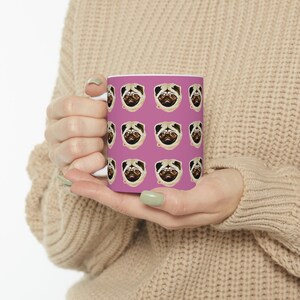 May include: A white ceramic mug with a pink background and a repeated pattern of pug faces. The pugs have brown eyes and black noses.