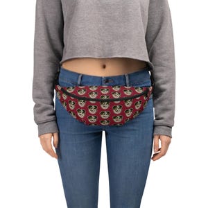 May include: A red fanny pack with a black adjustable strap and a repeating pattern of cartoon dog faces. The fanny pack is worn by a person wearing blue jeans and a grey cropped sweater.