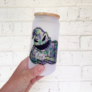 May include: A frosted glass tumbler with a bamboo lid. The tumbler features a colourful graphic of a character with a stitched appearance, holding dice. The character's design incorporates shades of green, purple, and black.