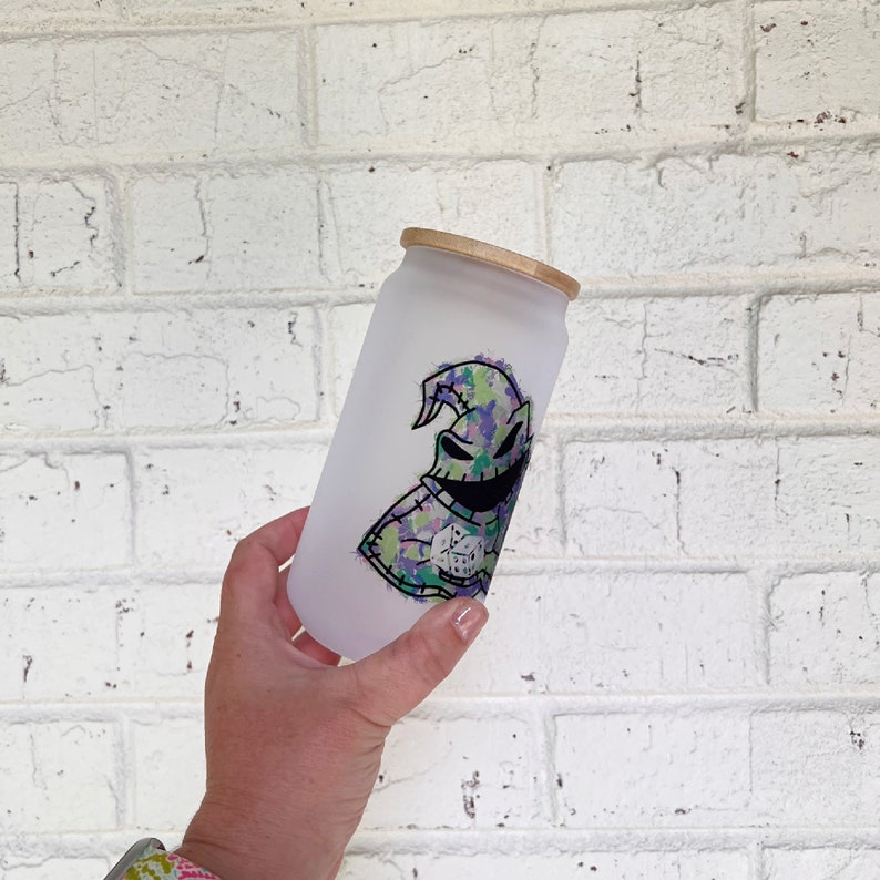 May include: A frosted glass can-shaped tumbler with a bamboo lid. The tumbler features a colourful graphic of a character with a stitched appearance, a wide grin, and a tall, pointed hat. The design incorporates shades of green, purple, and black.