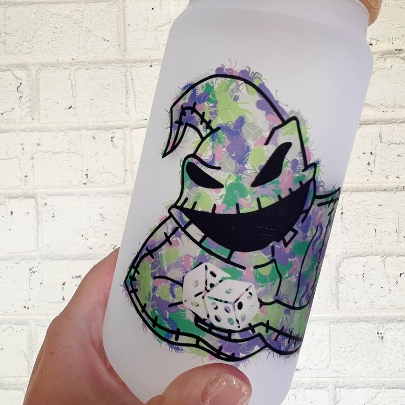 May include: A frosted glass water bottle with a colourful graphic of a stitched character with a wide grin and dice. The design incorporates green, purple, and white accents. The bottle has a wooden lid.