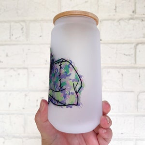May include: Frosted glass can-shaped tumbler with a bamboo lid. The tumbler features a colourful graphic of a monster with green, purple, and blue accents. The design is outlined in black. The tumbler is designed for cold beverages.