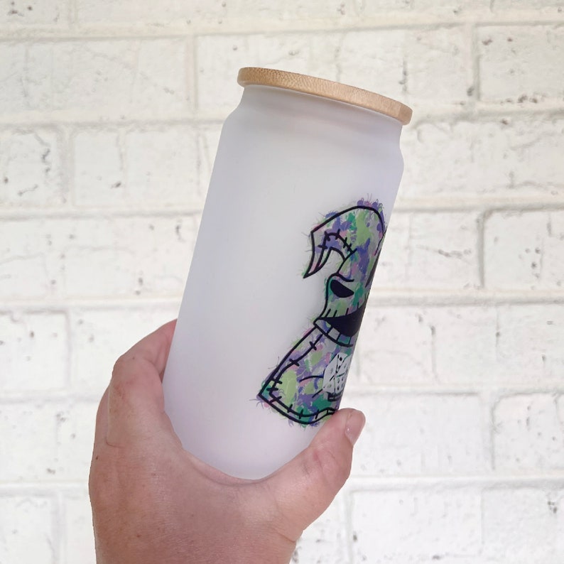May include: Frosted glass tumbler with a bamboo lid. The tumbler features a colourful graphic of a character with a stitched design in shades of green, purple, and black. The design is on a white background.