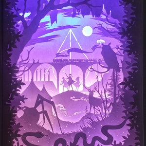 May include: A purple and blue paper cut-out art piece depicting a scene from the Harry Potter universe. The scene includes a train, a bridge, a forest, a deer, a snake, a spider, and three figures standing on a hill. The figures are holding wands and appear to be casting spells. The scene is illuminated from behind, creating a glowing effect.
