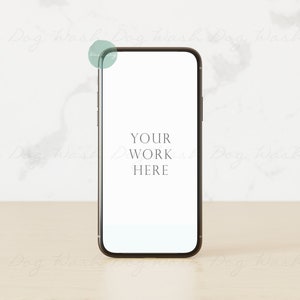 May include: A black smartphone mockup with a white screen displaying the text "YOUR WORK HERE" on a light brown wooden surface.
