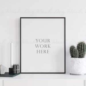 Puede incluir: Un póster enmarcado en negro con el texto "YOUR WORK HERE" sobre un fondo blanco. El póster está en un estante blanco con una vela blanca, lápices negros y un objeto geométrico gris sobre una superficie verde delante de él. Un cactus en una maceta blanca está en el lado derecho del estante.