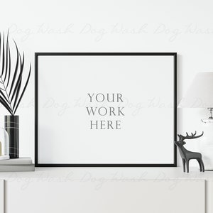 May include: A black framed picture mockup with the text "YOUR WORK HERE" on a white background. The picture is on a white shelf with a black and white vase, a glass cloche with a gold letter "a", and a white lamp with a white lampshade. A black reindeer figurine is on the shelf in front of the lamp.