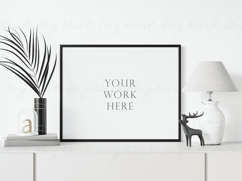 May include: A black framed picture mockup with the text "YOUR WORK HERE" on a white background. The picture is on a white shelf with a black and white vase, a glass cloche with a gold letter "a", and a white lamp with a white lampshade. A black reindeer figurine is on the shelf in front of the lamp.