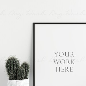 K&ouml;nnte beinhalten: Ein gerahmter Druck mit dem Text "YOUR WORK HERE" in minimalistischem Stil. Der schwarze Rahmen umgibt einen wei&szlig;en Hintergrund. Ein Topf-Kaktus mit gr&uuml;nen, stacheligen Bl&auml;ttern steht vor dem Rahmen.