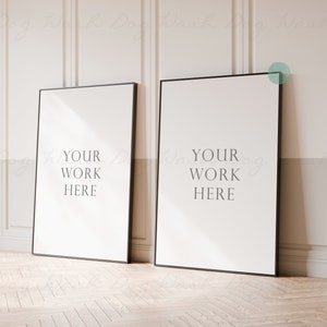 May include: Two black framed posters with white backgrounds and the text "YOUR WORK HERE" on each poster. The posters are leaning against a white wall with wainscoting.