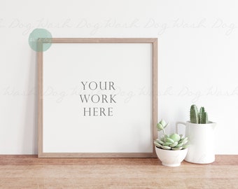May include: A light wood frame with a white mount and a white background. The text "YOUR WORK HERE" is printed in grey on the white background. The frame is sitting on a light wood surface. There are two small potted plants in front of the frame.