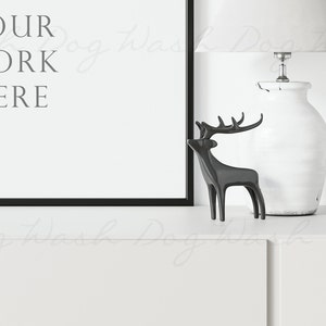 May include: A black framed picture with the text "YOUR WORK HERE" on a white background. A white ceramic lamp with a white lampshade is on the right side of the image. A black metal deer figurine is in front of the lamp. The picture, lamp, and deer are on a white surface.