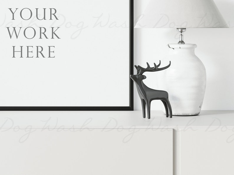 May include: A black framed picture with the text "YOUR WORK HERE" on a white background. A white ceramic lamp with a white lampshade is on the right side of the image. A black metal deer figurine is in front of the lamp. The picture, lamp, and deer are on a white surface.