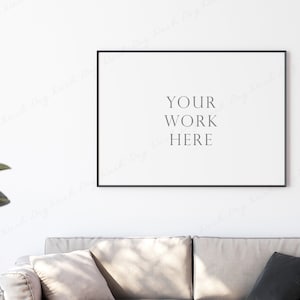 以下が含まれることがあります： 白い壁に「YOUR WORK HERE」という文字が書かれた黒い額縁の絵が、ベージュのソファの上に2つのクッションを置いて飾られています。