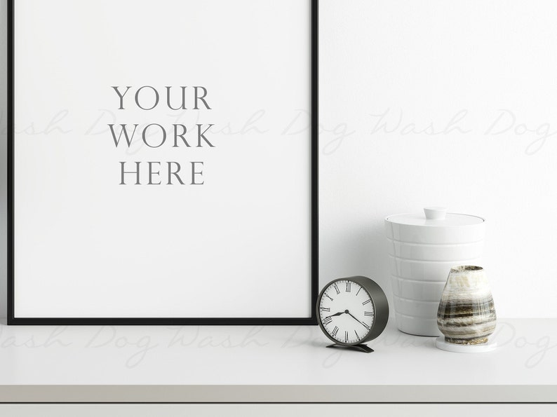 K&ouml;nnte beinhalten: Eine minimalistische Innenszene mit einem gerahmten Kunstwerk mit dem Text "YOUR WORK HERE". Eine kleine, runde Uhr, ein wei&szlig;er Beh&auml;lter und eine dekorative Vase mit Streifenmuster sind auf einer wei&szlig;en Oberfl&auml;che angeordnet.