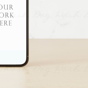 May include: A black smartphone with a white screen displaying the text "YOUR WORK HERE" is sitting on a light brown wooden surface.