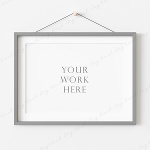 May include: A gray picture frame with a white mat hanging on a white wall. The frame is attached to a string with a wooden knob. The text "YOUR WORK HERE" is printed in the center of the mat.