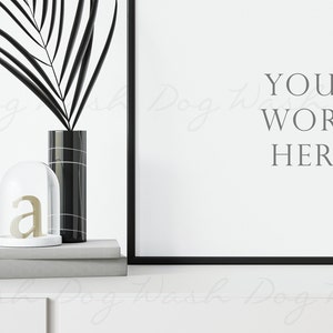 May include: A black and white framed poster mockup with the text "YOUR WORK HERE" on a white background. A black vase with a palm leaf and a glass cloche with a gold letter "a" are on a gray shelf in front of the poster.