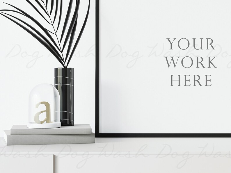 May include: A black and white framed poster mockup with the text "YOUR WORK HERE" on a white background. A black vase with a palm leaf and a glass cloche with a gold letter "a" are on a gray shelf in front of the poster.
