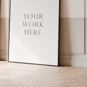 May include: Two black framed posters with white backgrounds are leaning against a white wall with decorative molding. The poster on the right has the text "YOUR WORK HERE" printed on it.