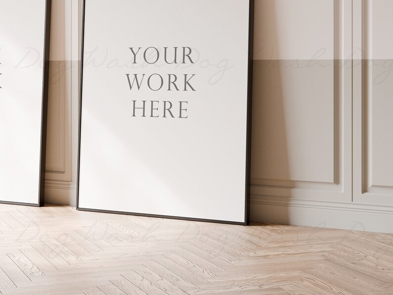 May include: Two black framed posters with white backgrounds are leaning against a white wall with decorative molding. The poster on the right has the text "YOUR WORK HERE" printed on it.