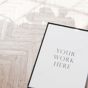 May include: A black picture frame with a white background and the text "YOUR WORK HERE" in the center. The frame is sitting on a light brown wood floor.