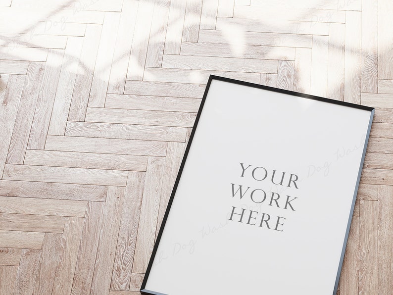 May include: A black picture frame with a white background and the text "YOUR WORK HERE" in the center. The frame is sitting on a light brown wood floor.