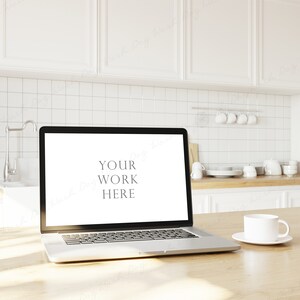 May include: A silver laptop computer with a blank white screen displaying the text "YOUR WORK HERE" is on a light wood table in a kitchen. A white coffee mug is on a white saucer to the right of the laptop.