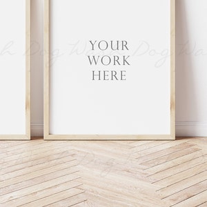 May include: Two wooden framed posters with white backgrounds, leaning against a white wall. The poster on the right has the text "YOUR WORK HERE" printed on it.