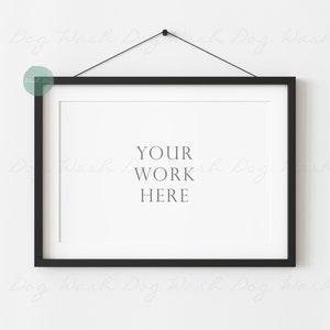 May include: A black picture frame with a white mount hanging on a white wall. The frame is hanging from a black cord with a black hook. The text "YOUR WORK HERE" is printed in the centre of the frame.