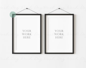 May include: Two vertical rectangular picture frames with thin black borders and white matting, each hanging from a black string. The center of each frame contains the white space with the centered black text: YOUR WORK HERE.
