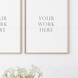 May include: Two framed prints with the text "YOUR WORK HERE" on a white wall with a small bouquet of white flowers in the foreground.