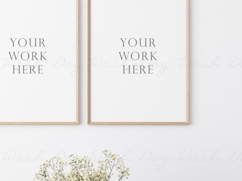 May include: Two framed prints with the text "YOUR WORK HERE" on a white wall with a small bouquet of white flowers in the foreground.