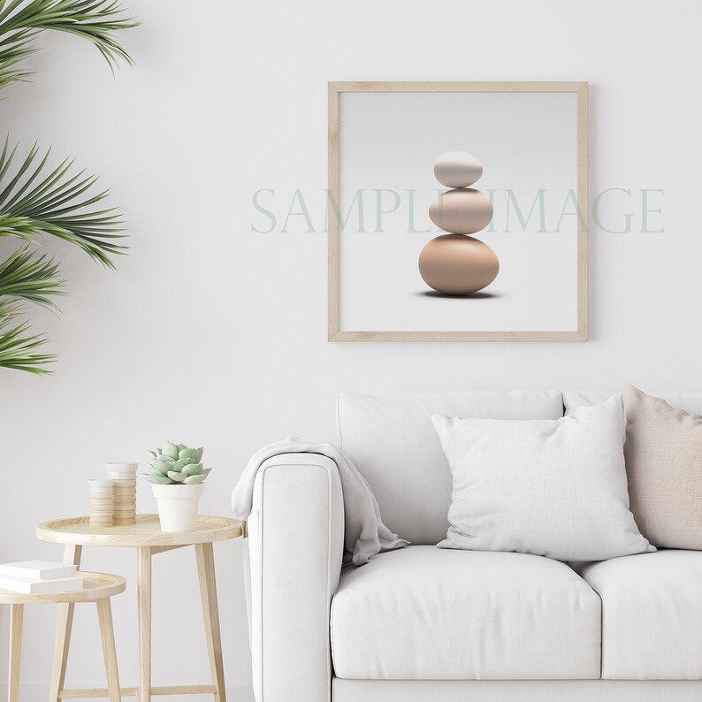 May include: A framed print of three stacked stones in shades of beige and white against a white background. The print is hanging on a white wall above a white couch with a throw blanket and two pillows.