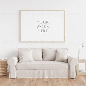 Puede incluir: Un sofá blanco con dos almohadas se encuentra frente a una pared blanca con un marco de madera alrededor de un póster blanco que dice "YOUR WORK HERE".