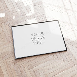 Op de afbeelding: Een zwarte gemonteerde foto mockup op een lichte houten vloer met de tekst "YOUR WORK HERE" in het midden van de lijst.