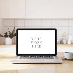 May include: A silver laptop computer with a white screen displaying the text "YOUR WORK HERE" is on a light wood table in a kitchen setting. A white ceramic mug is on a saucer to the right of the laptop.