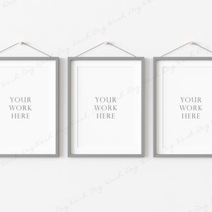 May include: Three framed art prints with gray frames and white matting are suspended from the wall. Each frame has the text "YOUR WORK HERE" in a simple font. The frames are hung by a black string and a small wooden peg.