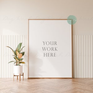 May include: A white frame with a wood border sits against a white wall with vertical grooves. The frame is empty with the text "YOUR WORK HERE" in the center. A potted plant with green leaves and a white pot sits on a wooden floor with a herringbone pattern.