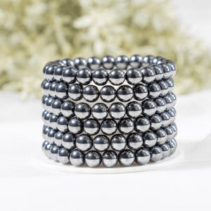 Könnte beinhalten: Ein Stapel von sechs Armbändern mit Hämatitperlen. Die Perlen sind dunkelgrau-metallisch und kreisförmig angeordnet. Die Armbänder werden auf einer weißen Oberfläche mit einem unscharfen Hintergrund präsentiert.